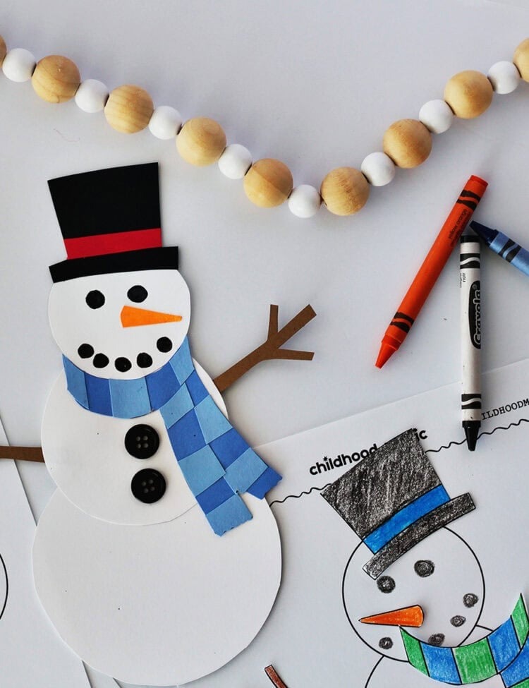 A snowman cut out of white cardstock with a blue scarf from cardstock. Next to the snowman are green, black, orange and blue crayons and a snowman printable with pieces that have been colored in.