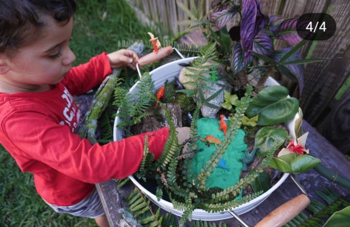 A child plays with a miniature garden setup, featuring toy dinosaurs, ferns, and other greenery in a bowl.