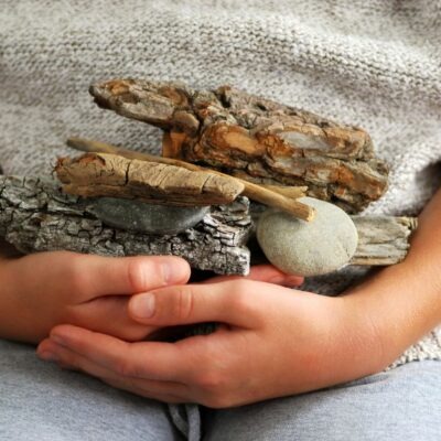 child holding a pile of bark, twigs, and rocks cupped in their hands - wearing a grey sweater and light colored blue jeans
