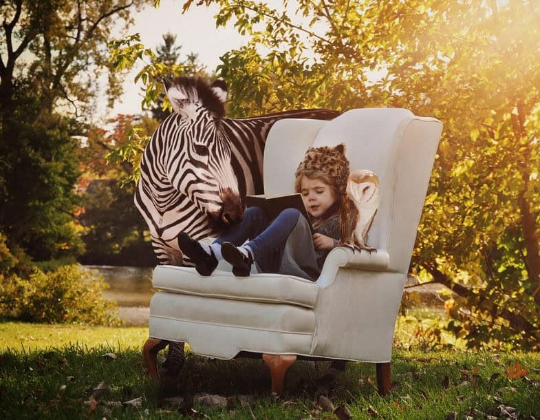 a young girl sitting in a large arm chair outside reading a book with a zebra looking over her shoulder