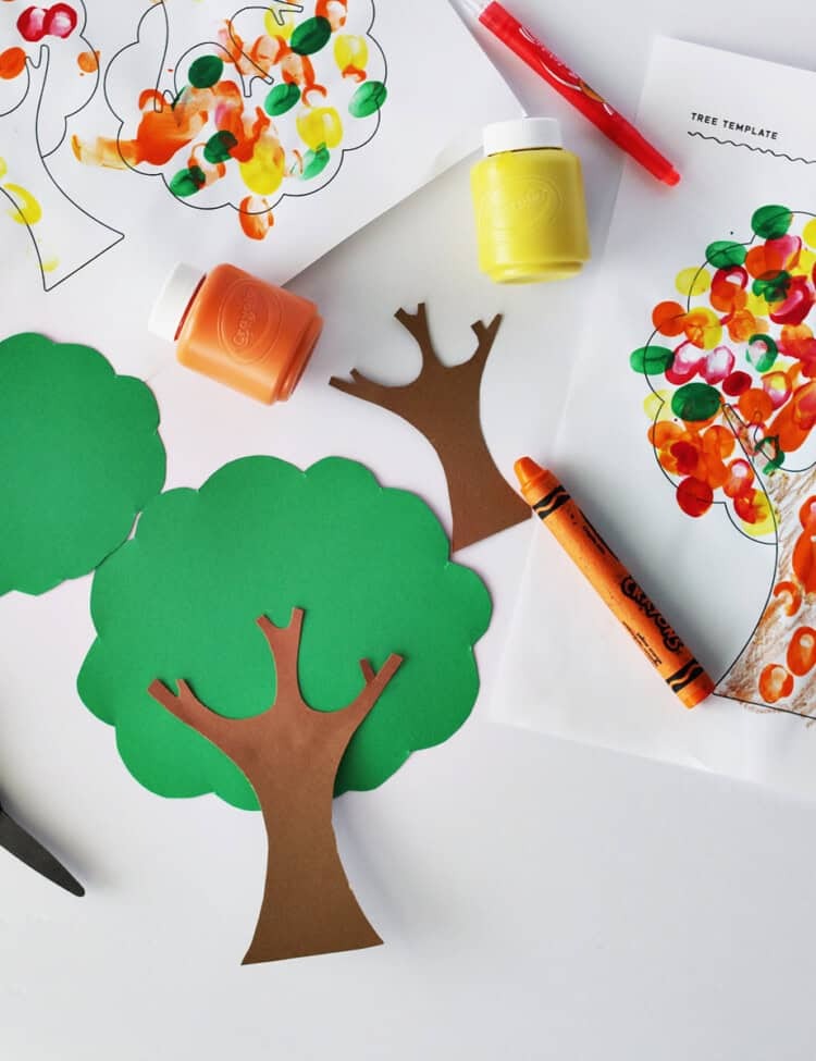 tree cut out of brown and green cardstock next to kids scissors, crayons, and a finger painted tree coloring page on a white table.
