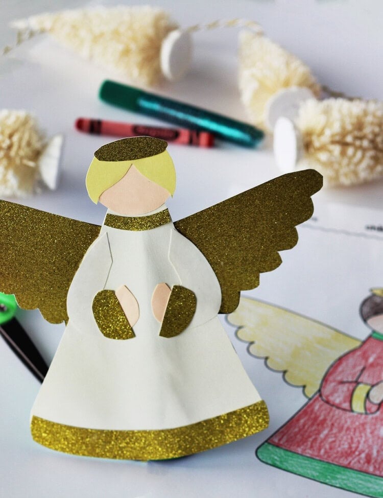 Angel tree topper made from cardstock next to an angel coloring page on a table with kids art supplies and a Christmas tree garland.